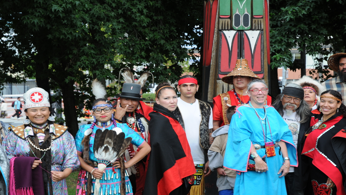 Group of people dressed in traditional native american clothing, with a Totem pole in the middle